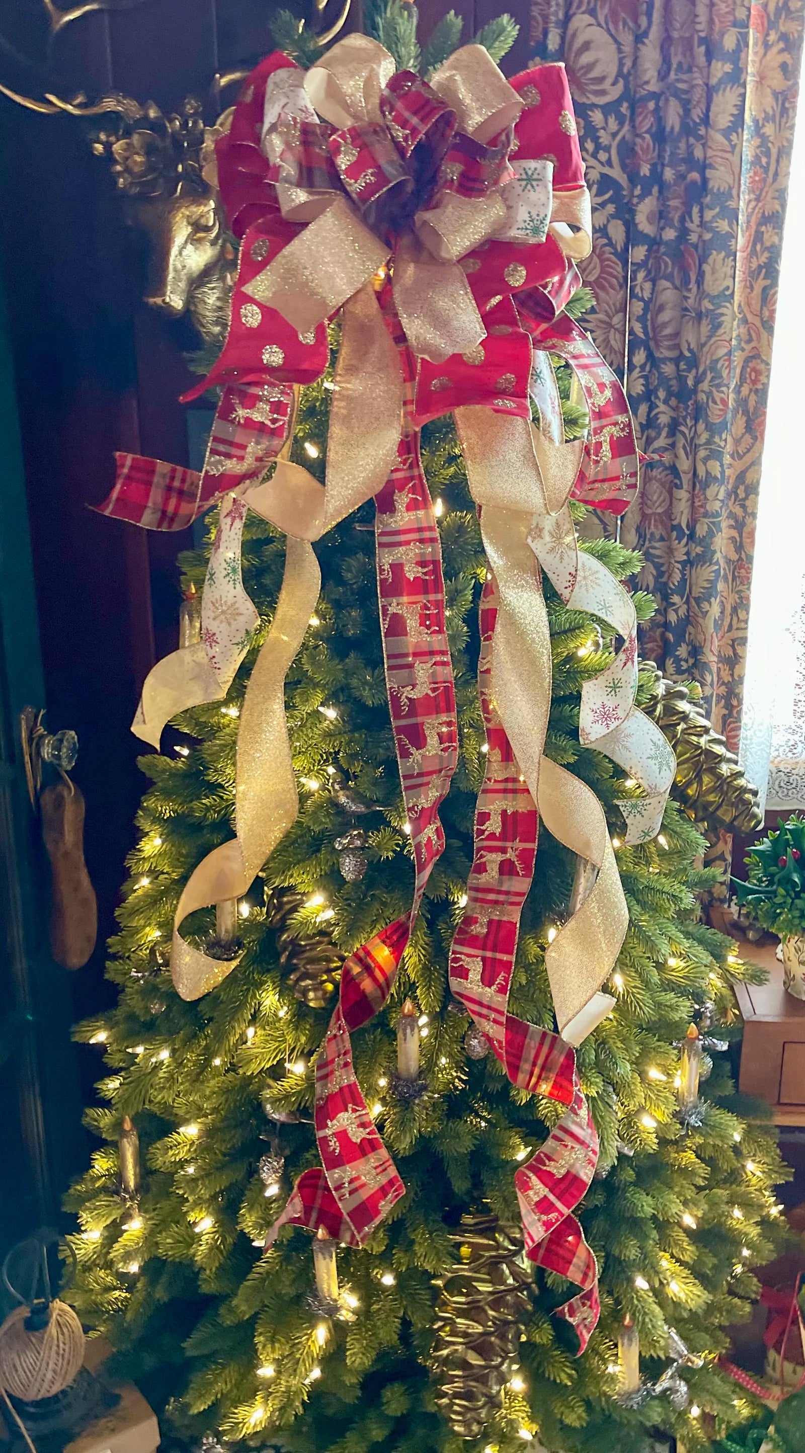 May include: A green Christmas tree decorated with a large gold and red bow, gold and red plaid ribbon, and white ribbon with snowflakes. The tree is also decorated with white lights.