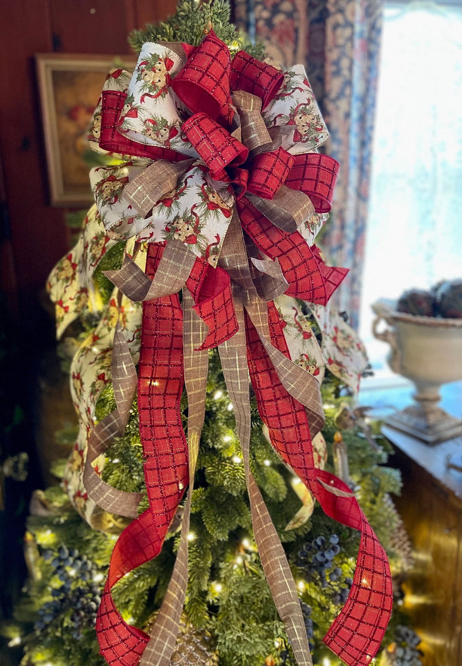 May include: Christmas tree topper bow in red and beige plaid ribbon. The bow features a burlap-like texture and glittery accents.  The ribbons are arranged in a cascading style, creating a lush and festive look. The design incorporates a cream-colored ribbon with a vintage-style Christmas print. This decorative tree topper is perfect for adding a touch of rustic elegance to any Christmas tree.