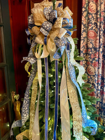 May include: A decorative Christmas tree topper bow featuring multiple loops and long cascading streamers. The ribbons are crafted in a combination of metallic gold, solid navy blue, and light blue fabric with a white floral pattern throughout.