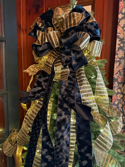 May include: A decorative tree topper bow featuring a combination of black velvet ribbons and metallic gold striped ribbons. The bow has a large, multi-looped center with long, cascading ribbon tails that drape downward over the evergreen branches.