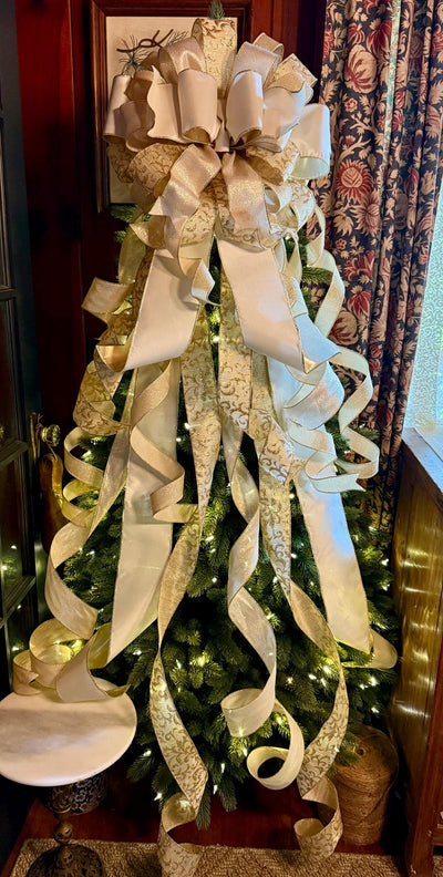 May include: A large decorative Christmas tree topper bow featuring multiple loops and long, cascading ribbon tails. The design combines solid cream-colored fabric with shimmering gold metallic patterned ribbon, creating a layered effect.