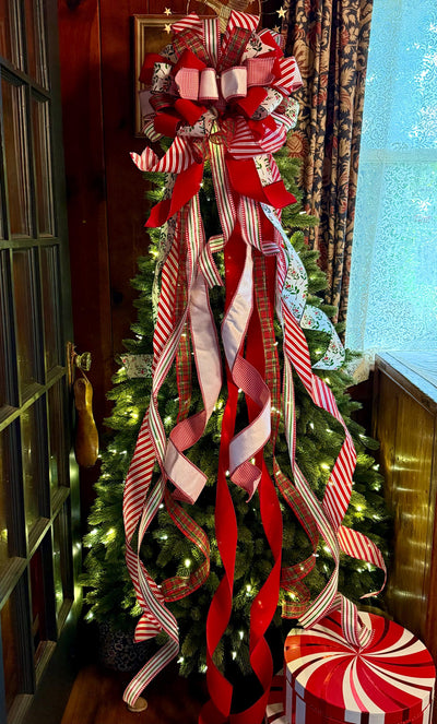 May include: A large Christmas tree topper bow featuring multiple long, cascading ribbons in red, white, and green patterns, including stripes, plaid, and holly prints, attached to the top of a lit evergreen tree with a round peppermint gift box.
