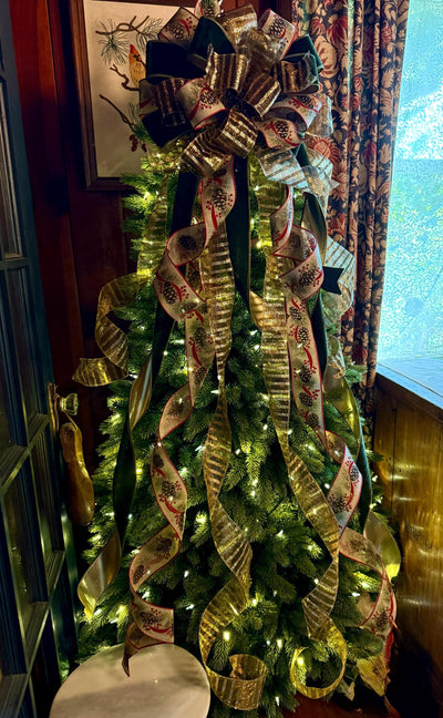 May include: A Christmas tree topper featuring a large, multi-layered bow made of dark green velvet, gold metallic striped, and cream-colored ribbon with a pinecone and red berry print. Long, cascading ribbon tails drape down the evergreen branches.