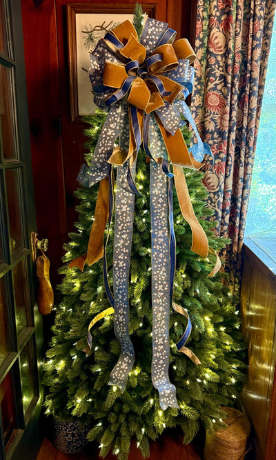 May include: A large decorative Christmas tree topper bow featuring multiple loops and long cascading tails. The bow is crafted from a combination of blue fabric with a white floral pattern and solid gold-toned velvet ribbon, creating a layered look.